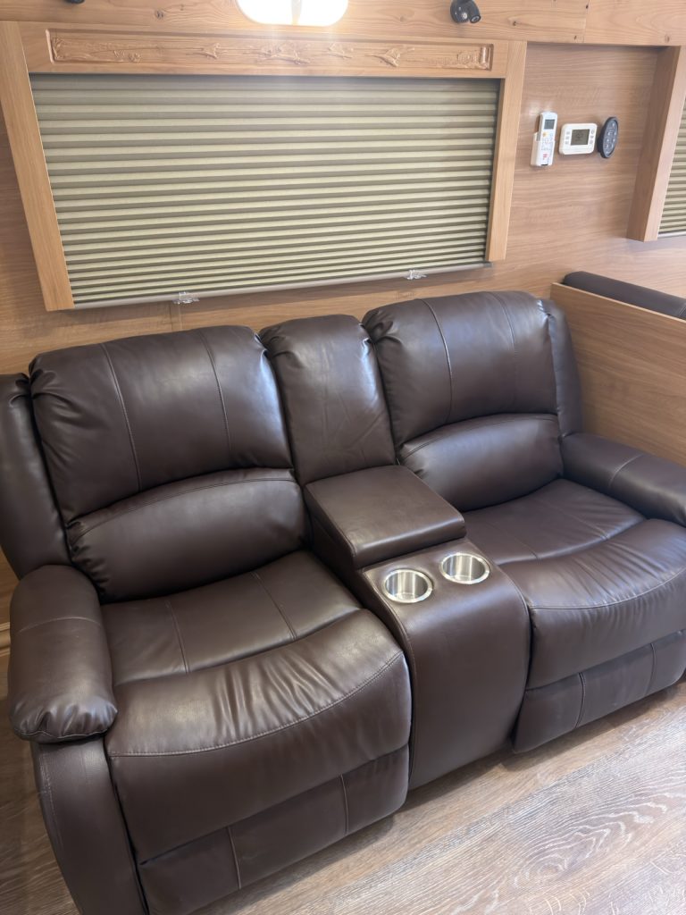 Brown two-person recliner with center cupholder console beneath a shaded window.