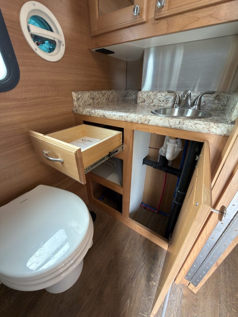 Bathroom vanity with stainless sink, open shelf, drawer, and exposed plumbing.