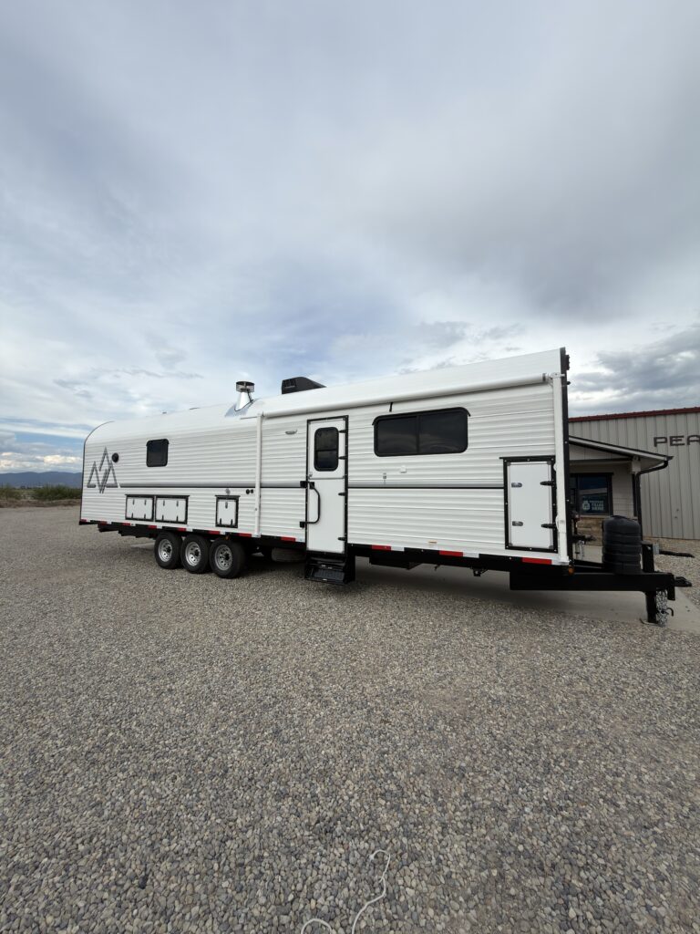 Side exterior view of the white Schrauth Camp showing the awning, entry door, storage compartments, and triple-axle chassis