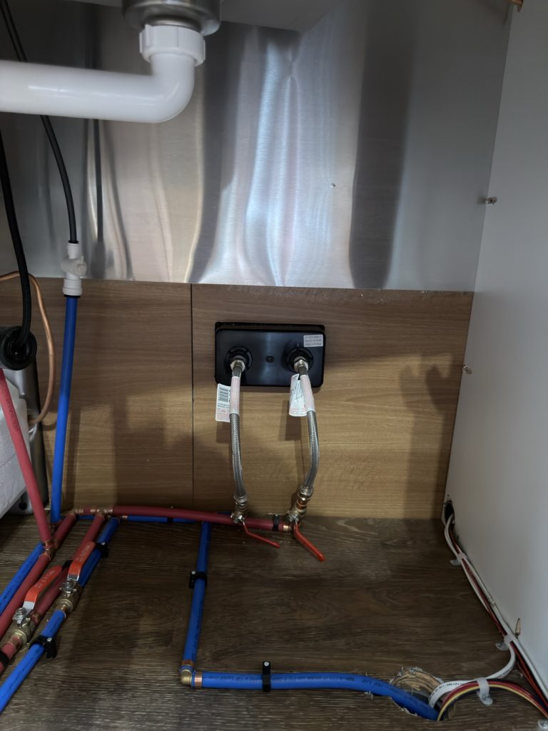 Exposed red and blue water lines, drain, and outlets beneath the kitchen cabinetry.