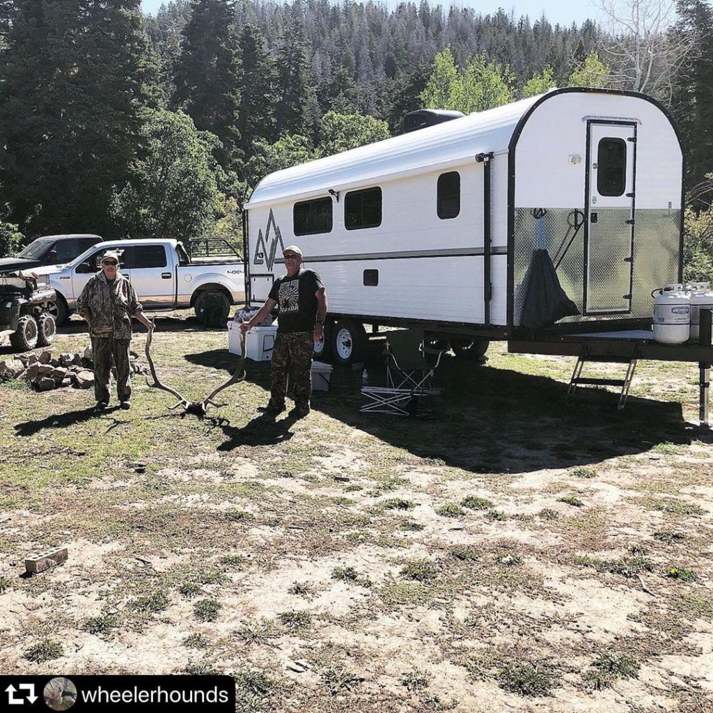 Hunters gathered beside a Peak camp trailer at a wooded campsite