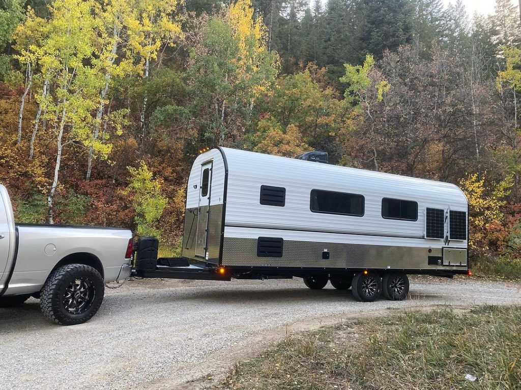 Pickup towing a long custom Peak camp trailer through a forest campsite
