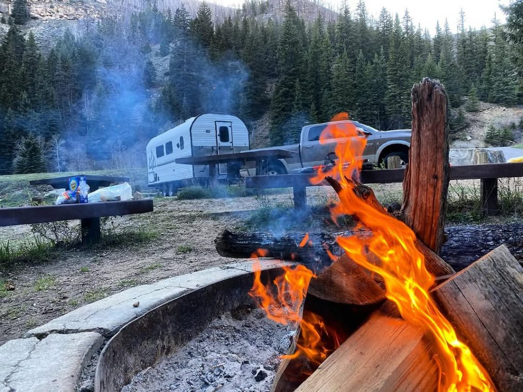 Bright campsite fire in the foreground with a Peak camp trailer among mountain trees
