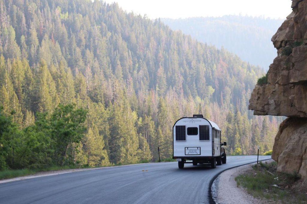 Rear view of a Peak camp trailer rounding a mountain road above a forested valley