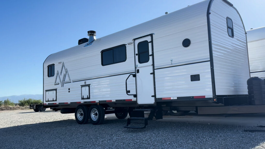 White Plankis Camp #124 with black trim, side entry steps, tandem wheels, and mountain graphics on a gravel lot.