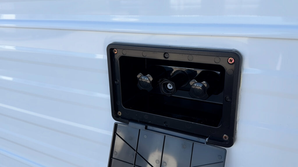 Exterior shower and spray port with two control knobs and a hose connection behind an open cover on Plankis Camp #124.