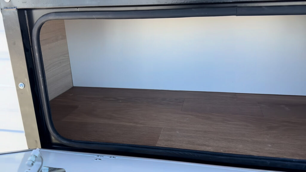 Large open exterior storage compartment with white walls, a wood-look floor, and a black seal on Plankis Camp #124.