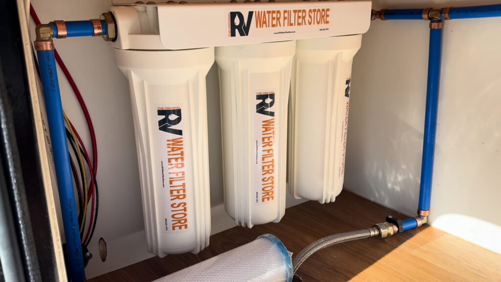 Three white water filter housings mounted beside blue water lines inside a compartment on Plankis Camp #124.