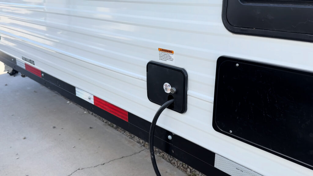 Power cord plugged into a black exterior inlet beside a service panel on Plankis Camp #124.