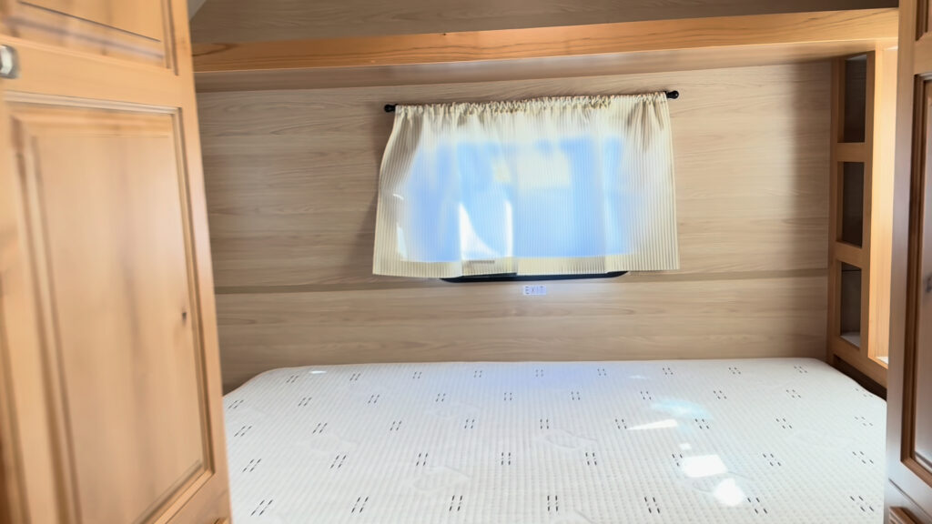 Bed beneath a curtained window in Plankis Camp #124, framed by tall wood cabinets and open side shelving.