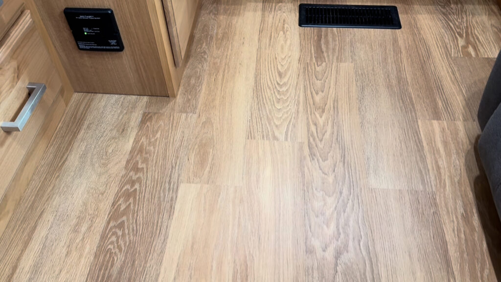 Wood-look plank flooring in the aisle of Plankis Camp #124, beside cabinetry and a black floor vent.