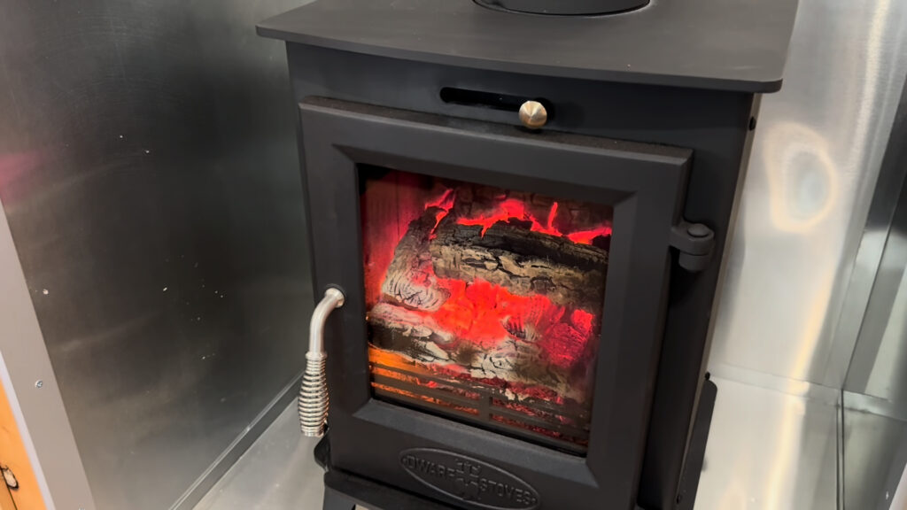 Black glass-front stove with glowing logs and metal wall shielding inside Plankis Camp #124.