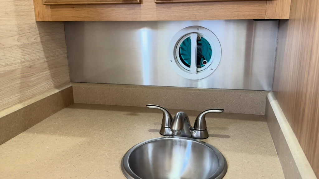 Bathroom sink and faucet beneath a mirrored cabinet, with a round wall vent behind them in Plankis Camp #124.