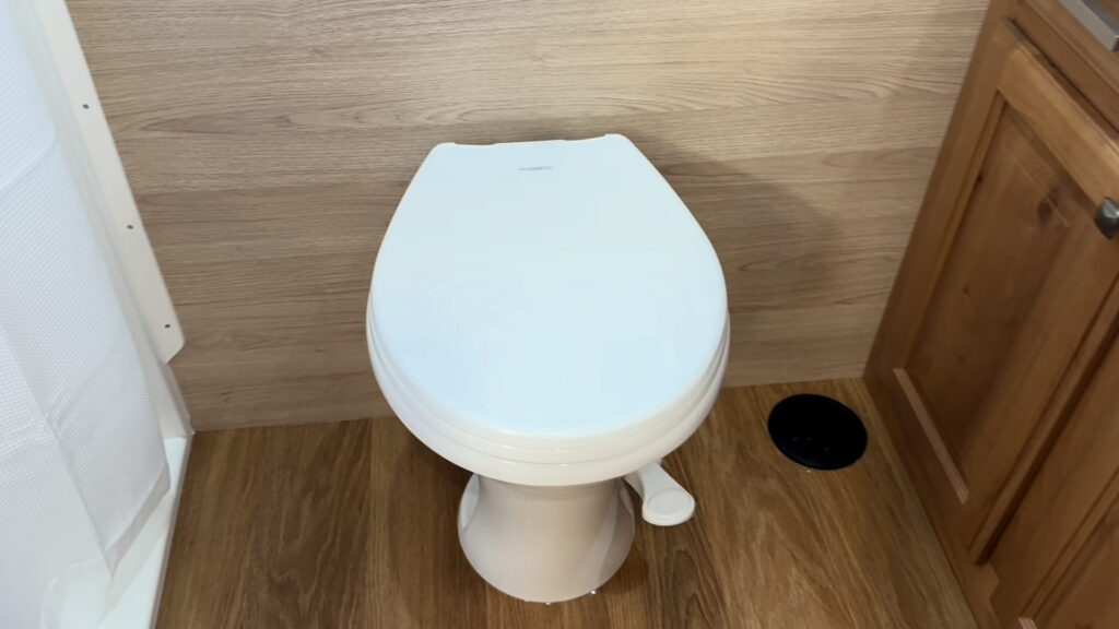 White toilet beside a wood cabinet and dark floor opening inside the bathroom of Plankis Camp #124.