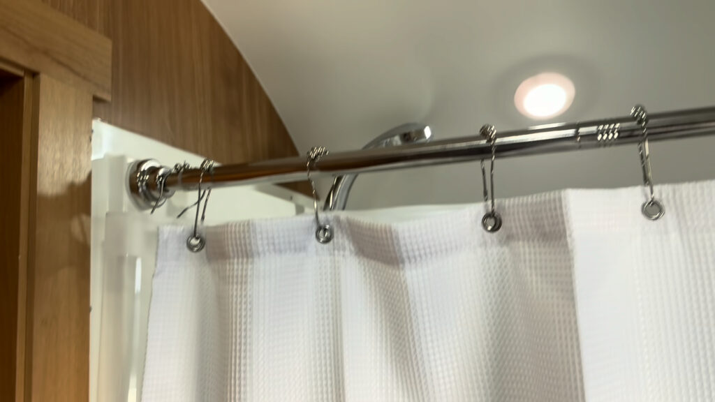 Metal shower curtain rod with rings and a white curtain below a ceiling light in Plankis Camp #124.