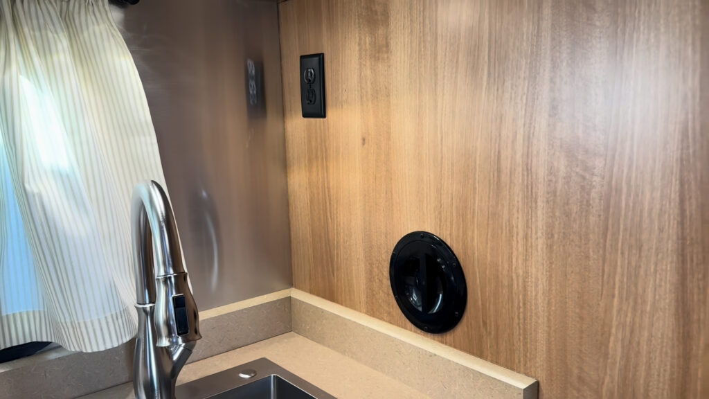 Black power outlet and round shower plumbing access port beside the kitchen faucet in Plankis Camp #124.