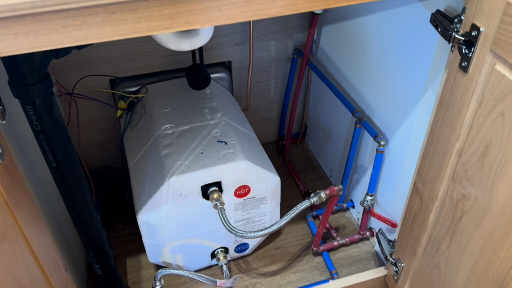 White water heater, red and blue water lines, and black drain plumbing under the kitchen sink in Plankis Camp #124.