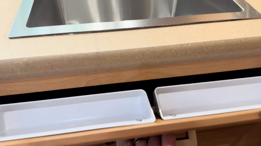 Two white tip-out storage trays opened beneath the stainless kitchen sink in Plankis Camp #124.