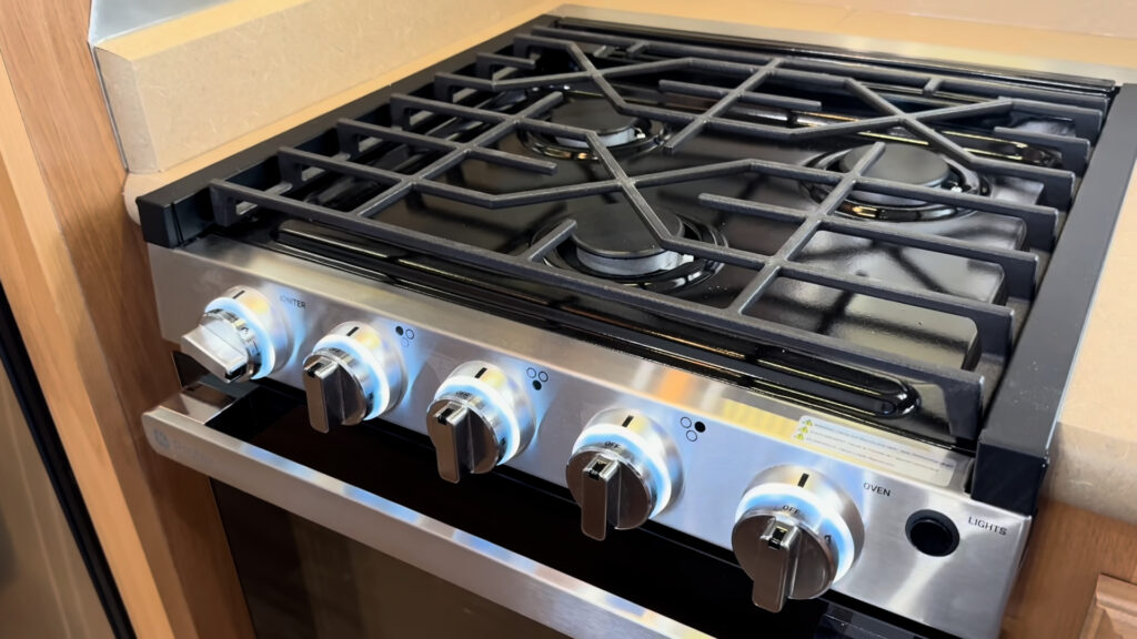Black cooktop grates, burners, and a row of metal control knobs on the kitchen range in Plankis Camp #124.