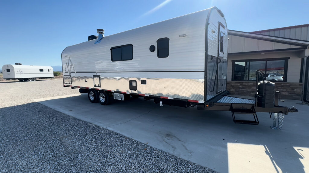 Side view of the Kendall Camp showing its long white exterior, tandem wheels, front entry, and diamond-plate lower panels.