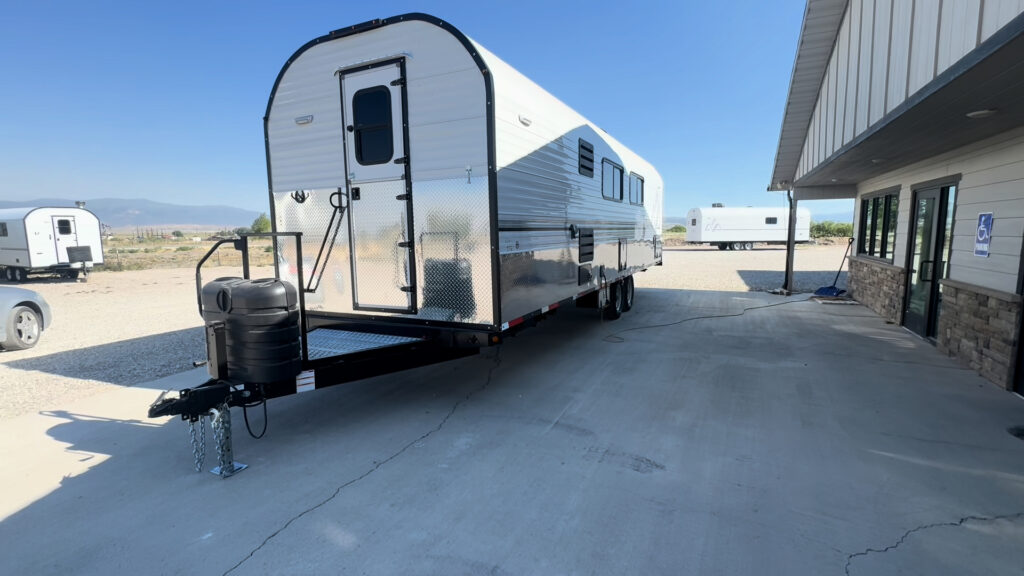 Front three-quarter view of the Kendall Camp showing the entry door, fold-up steps, tongue deck, and propane cover.