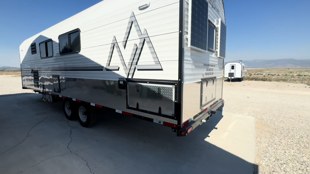 Rear side view of the Kendall Camp showing tandem wheels, windows, and diamond-plate storage doors.