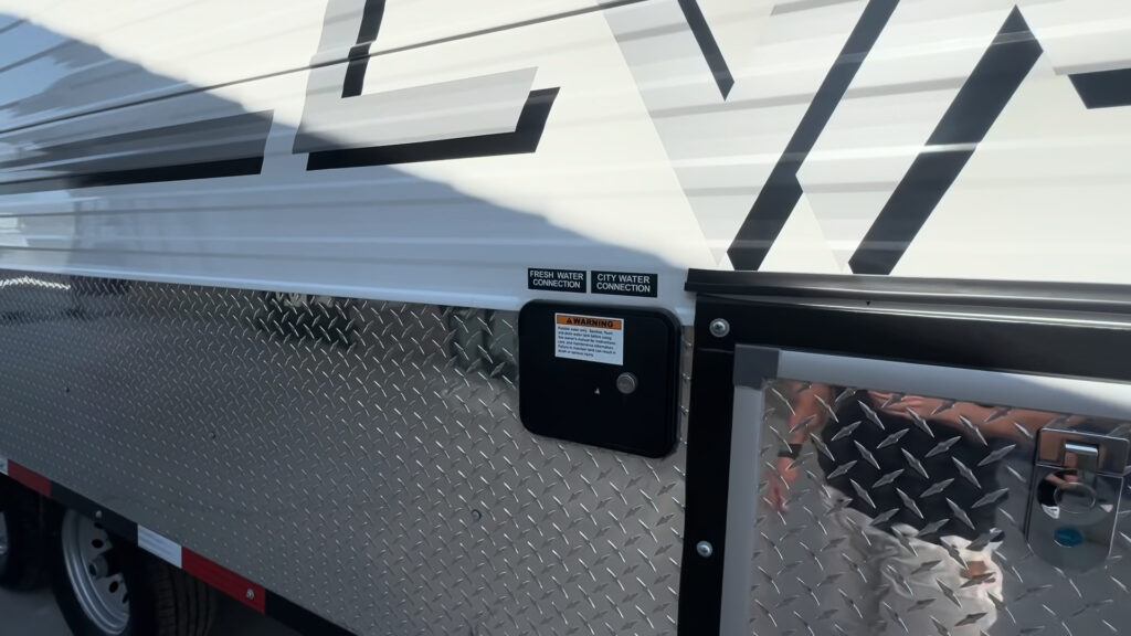 Close view of an exterior utility connection panel beside a diamond-plate compartment door on the Kendall Camp.