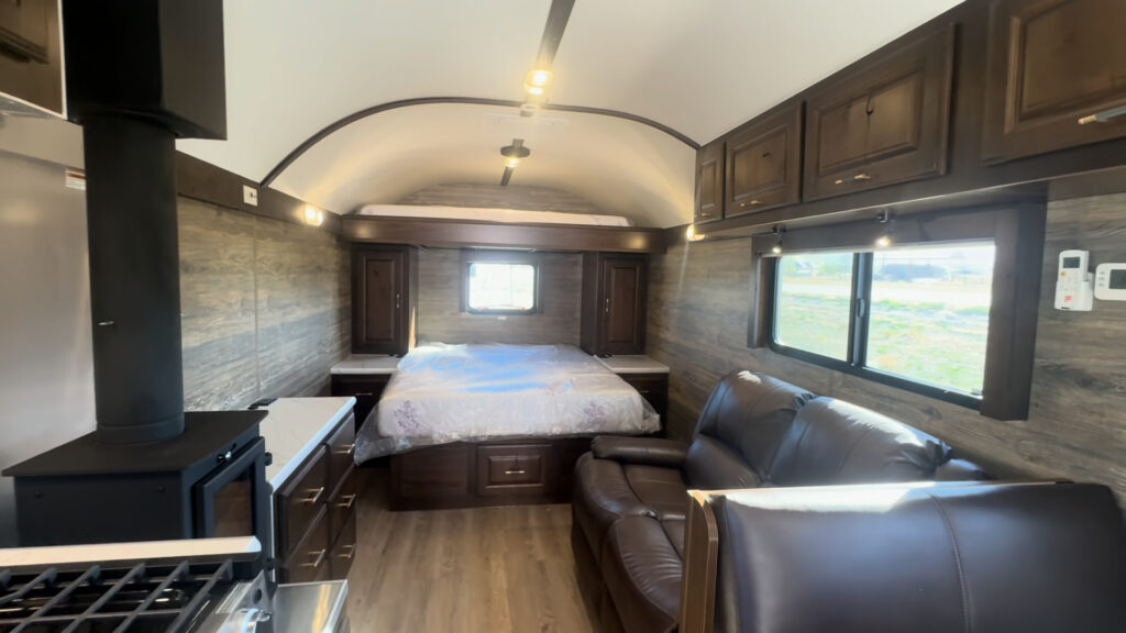 Wide interior view of the Kendall Camp cooktop, wood-burning stove, reclining sofa, and queen bed.