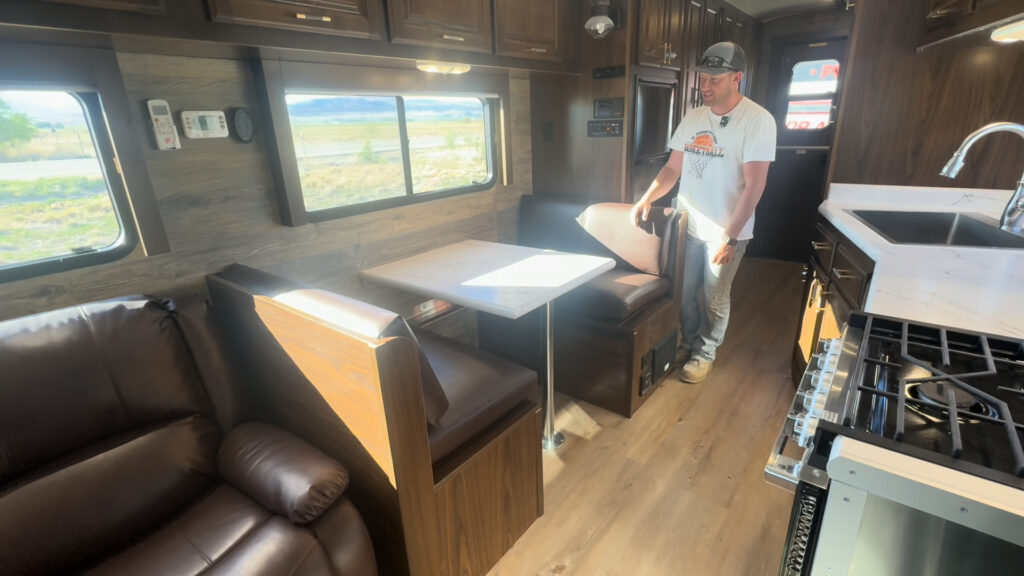 Interior view of the Kendall Camp dinette, reclining sofa, side windows, and kitchen, with a person standing beside the table.