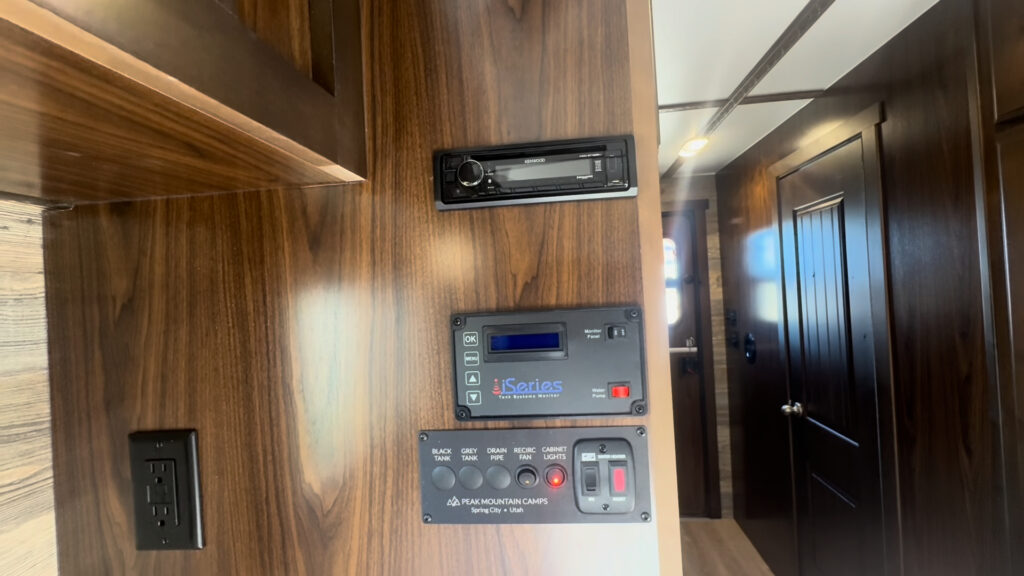 Close view of the Kendall Camp stereo, tank monitor, switches, and outlet on a wood-paneled wall.
