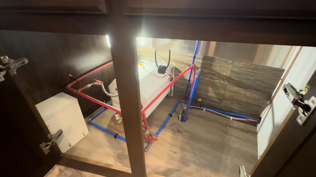 Open Kendall Camp cabinet showing water lines and plumbing connections beneath a counter.