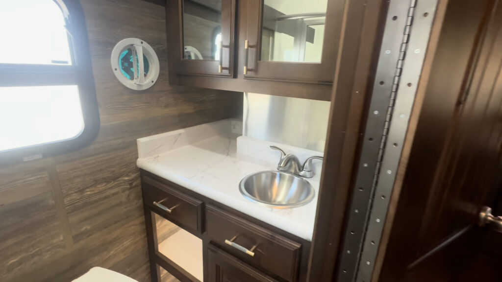 Interior view of the Kendall Camp bathroom vanity with a small stainless steel sink, mirror, hardwood cabinets, and frosted window.