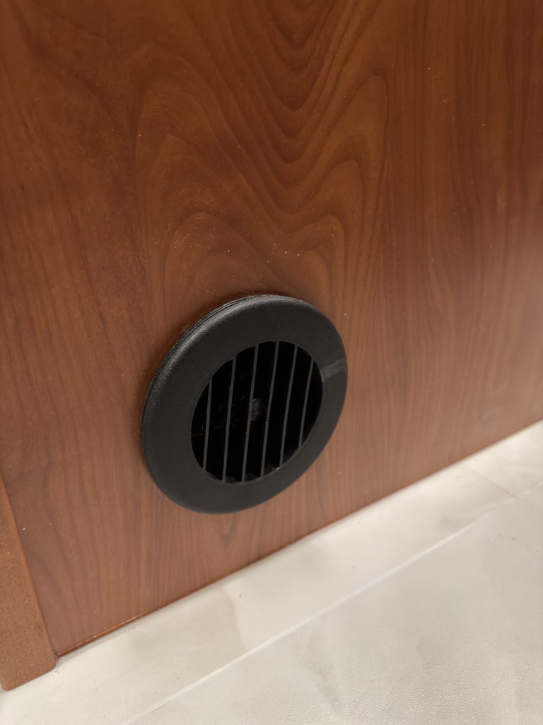 Round black vent grille in a wood cabinet base in Keulen Camp #132.