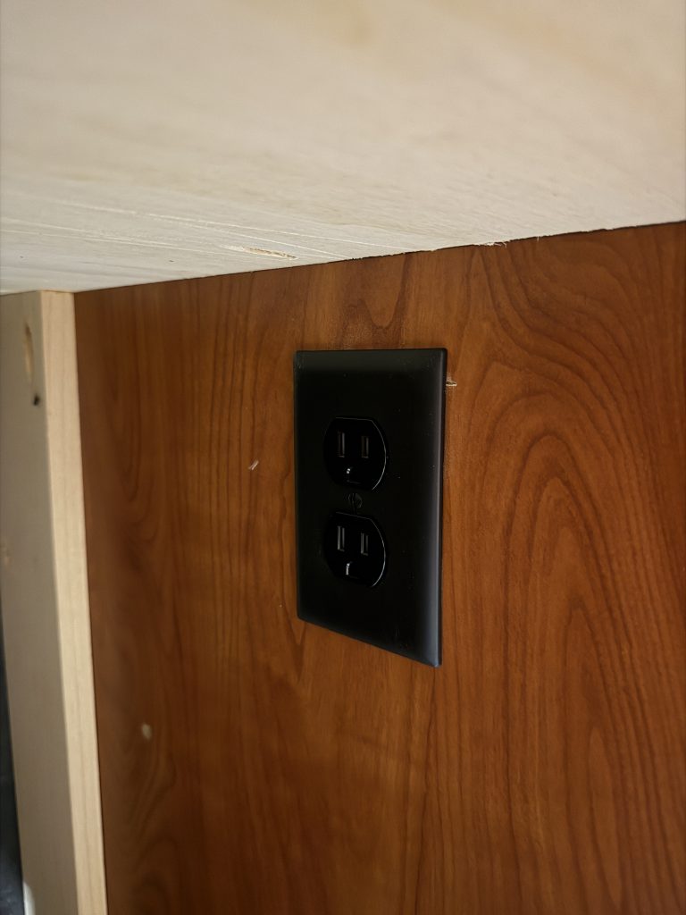 Black duplex power outlet in wood paneling beside the office desk in Keulen Camp #132.