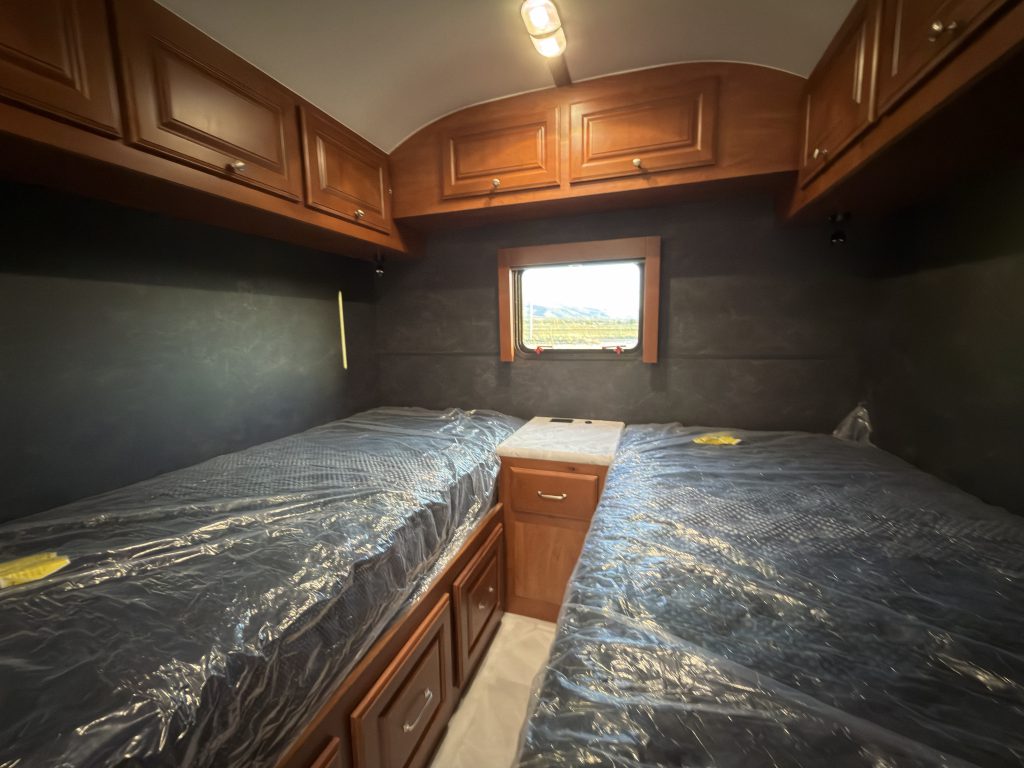 Twin mattress platforms with a central nightstand beneath overhead cabinets and an uncovered end window in Keulen Camp #132.
