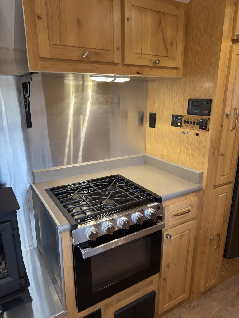 Van Niewkoop Camp #136: GE 3-burner cooktop and oven beneath a metal backsplash and upper wood cabinetry.