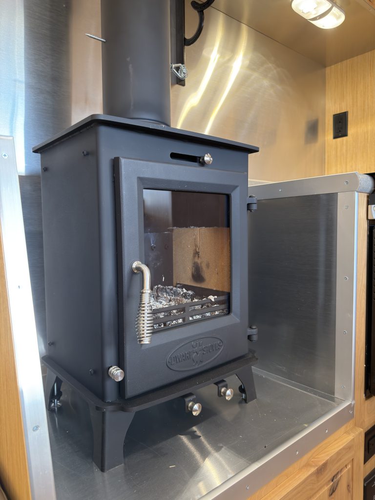 Van Niewkoop Camp #136: Close view of the small wood-burning stove with its glass-fronted door and flue.
