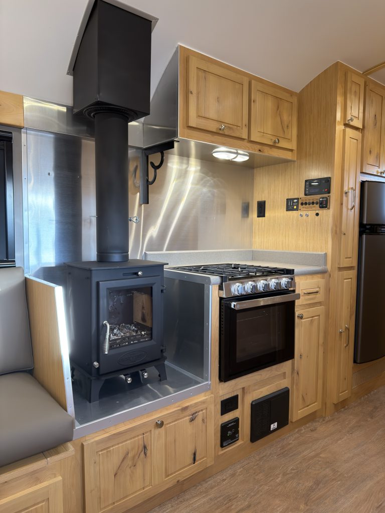 Van Niewkoop Camp #136: Kitchen appliance wall with the GE range, wood-burning stove, and hardwood upper cabinets.