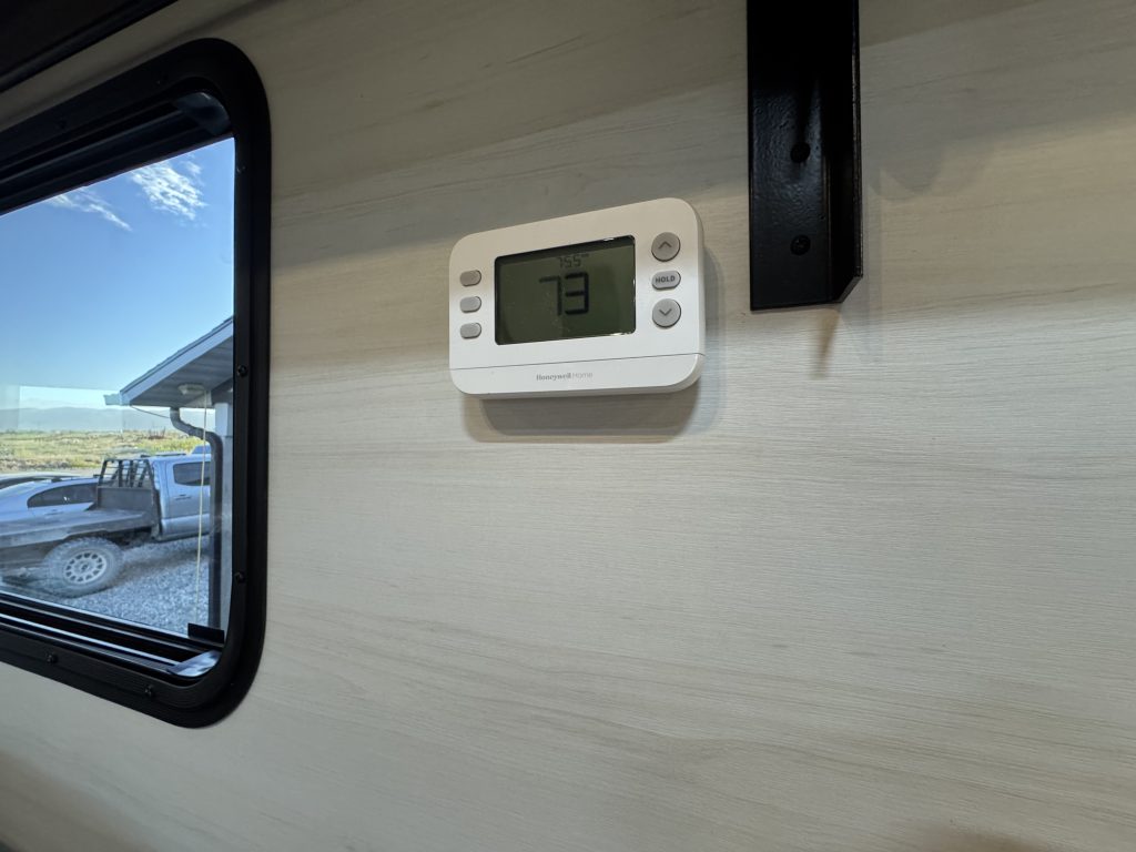 Wall-mounted digital thermostat beside a window and dark trim in Watson Camp #134.