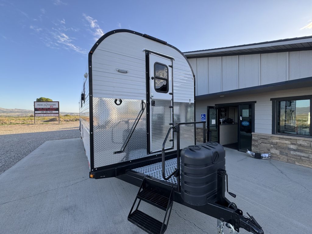 Curved front wall with entry door, steps, handrails, and trailer tongue with covered propane cylinders in Watson Camp #134.