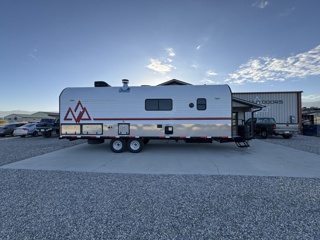 Full side view of the camp with windows, a Peak graphic, tandem wheels, and front entry in Watson Camp #134.