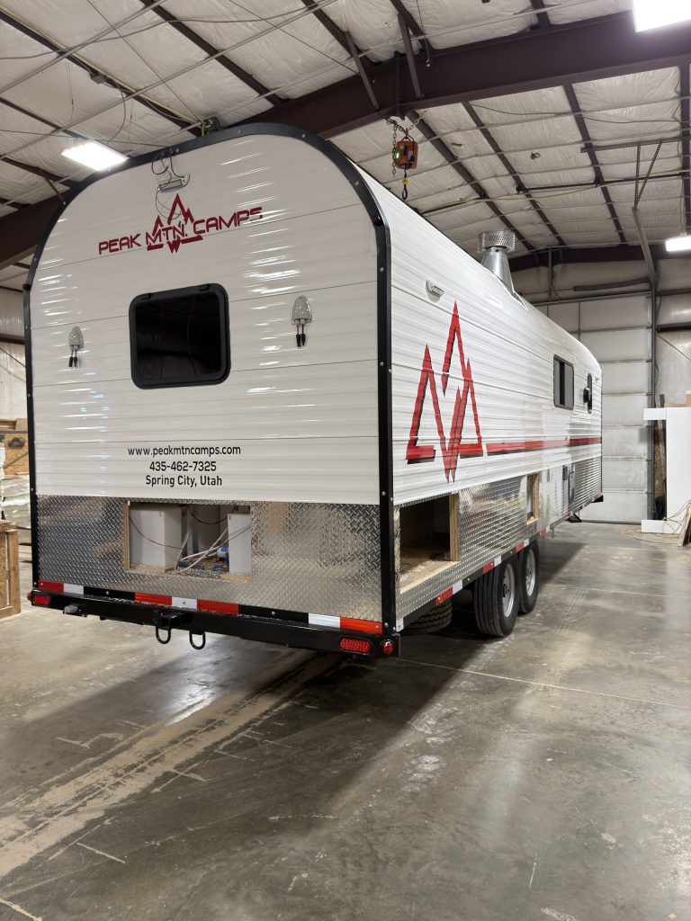 Rear and roadside view inside the workshop, showing white siding, red Peak graphics, tandem wheels, and open lower compartment bays in Watson Camp #134.