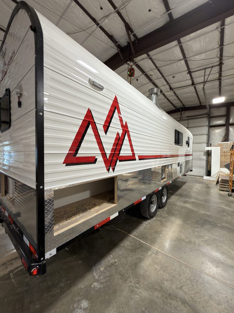 Close roadside angle inside the workshop, showing the red Peak graphic, tandem wheels, and open lower compartment bays in Watson Camp #134.