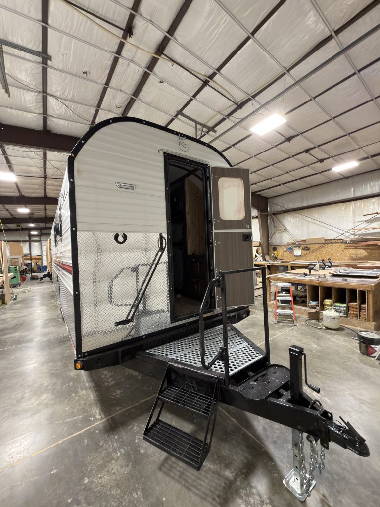 Front exterior inside the workshop, showing the open entry door, steps, handrails, front platform, and trailer hitch in Watson Camp #134.